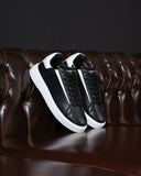 A pair of black and white low-top sneakers with white platform soles, displayed on a dark brown tufted leather sofa against a dark background.