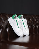 A pair of white low-top sneakers with vibrant green accents on the heel tab and side stripes, displayed on a dark brown tufted leather sofa against a dark background.