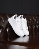 A pair of white leather low-top sneakers with white laces and thick white soles, displayed on a dark brown tufted leather sofa against a dark background.