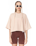 A model wearing a light beige oversized short-sleeve crop top and dark brown bike shorts, styled with white-framed sunglasses, against a plain white background.