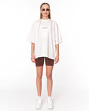 A model stands against a white studio background wearing an oversized white t-shirt with 'johnnie life' printed in black on the chest, brown fitted shorts, white sneakers, and sunglasses.