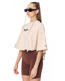 A woman modeling a beige cropped t-shirt with black graphic text on the chest, paired with brown fitted shorts, against a plain white background.