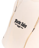 Close-up of a light cream or off-white garment with black gothic-style 'Martin Valen' text printed on the chest, with thin gold-toned braided cords hanging near the shoulder area.