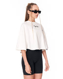 A model wearing a white cropped tunic top with a black script logo on the chest, paired with black bike shorts and black sunglasses with long chains, against a plain white background.
