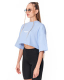 A woman modeling a light blue cropped t-shirt with a white chest graphic, paired with black biker shorts, against a plain white background.
