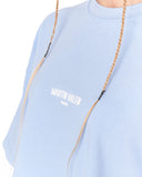 A close-up of a person wearing a light blue crewneck t-shirt with 'MARTIN VALEN' printed in white on the chest. The person has blonde braided hair hanging down the front of the shirt, set against a plain white background.