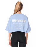 A woman viewed from the back wearing a light blue cropped T-shirt with white text reading 'MARTIN VALEN' and smaller script text below, paired with black leggings against a plain white background.