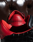 A close-up shot of a red leather shoe heel tab with 'MARTIN VALEN' embossed in black, resting on a black crocodile-embossed surface against a dark brown tufted leather background.