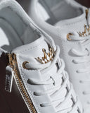 White leather high-top sneakers featuring gold crown-shaped eyelets and a gold side zipper, shown in a close-up shot.