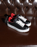 A pair of low-top sneakers with black croc-embossed leather uppers, white laces, and white rubber soles, featuring red lining and a black tongue with white text, displayed on a brown leather tufted sofa.