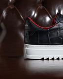 A close-up shot of a black leather sneaker with a textured crocodile pattern, a thick white platform sole, and a vibrant red inner lining, set against a dark brown tufted leather background.