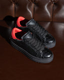 A pair of black low-top sneakers with a glossy crocodile-embossed texture, black laces, and a vibrant red interior lining, displayed on a dark brown leather tufted sofa.
