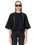 A model wearing a black oversized short-sleeve crop top with tonal graphic embroidery, paired with black leggings and black sunglasses, against a plain white background.