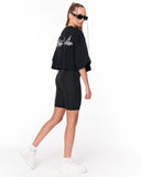 A woman modeling a black cropped t-shirt and matching black biker shorts set, shown from a rear three-quarter angle against a plain white studio background. The shirt features white script lettering on the back.