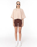A woman modeling a light beige short-sleeve cropped top and dark brown fitted bike shorts, styled with white sneakers and sunglasses against a plain white studio background.