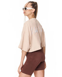 A woman shown from the back wearing a beige cropped t-shirt with white script lettering on the back, paired with brown bike shorts, against a plain white background.