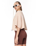 A woman shown in side profile wearing an oversized light beige cropped top, brown fitted biker shorts, and white sunglasses against a plain white studio background.