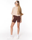 A woman modeling a beige cropped tunic top and dark brown fitted biker shorts with white sneakers against a plain white studio background.