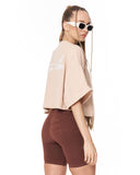 A woman shown from the back wearing a light beige oversized crop top with white script text on the back and dark brown fitted biker shorts, against a plain white background.