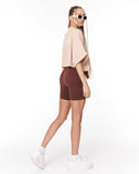 A woman modeling a beige oversized t-shirt and dark brown fitted bike shorts, paired with white sneakers and white-framed sunglasses, shown from a side-back angle against a plain white studio background.