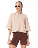 A model wearing a light beige oversized short-sleeve crop top and dark brown fitted bike shorts, styled with white sunglasses, against a plain white background.