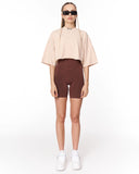 A model stands against a white studio background wearing a light beige cropped short-sleeve top, dark brown fitted biker shorts, white sneakers, and black sunglasses.