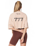 A woman shown from the back wearing a light peach-pink cropped t-shirt with a black '777' graphic and 'Martin Valen' signature, paired with brown bike shorts against a white background.