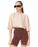 A woman modeling a light beige cropped t-shirt with wide short sleeves and a high neck, paired with dark brown biker shorts, against a plain white background.
