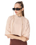 A model wearing a pale blush pink short-sleeve crop top with a high collar featuring small black geometric details, styled with black sunglasses and a brown skirt against a white background.