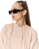 A model with a high ponytail wearing black square sunglasses with a gold chain strap hanging down her face, paired with a light pink sweatshirt against a white background.