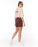 A model shown from the back wearing a light beige cropped t-shirt with black graphic text and dark brown fitted biker shorts, paired with white sneakers, against a plain white studio background.