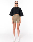 A model stands against a white studio background wearing a black cropped short-sleeve top, beige high-waisted biker shorts, white sneakers, and black sunglasses.