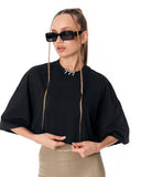 A woman modeling a black oversized short-sleeve top with a crew neck, paired with beige pants and black sunglasses with braided chains, against a plain white background.