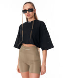 A model wearing a black cropped t-shirt with wide three-quarter sleeves and beige high-waisted bike shorts, styled with black sunglasses and a silver collar necklace against a plain white background.