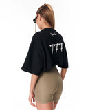 A woman shown from the back wearing a black cropped t-shirt with a large white '777' graphic and a small white script logo on the upper back, paired with beige fitted shorts against a plain white background.