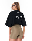 A woman shown from the back wearing a black oversized cropped T-shirt with a large white '777' graphic and a small white script logo on the neck, paired with olive green fitted shorts against a plain white background.