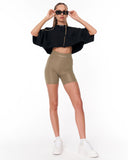 A model wearing a black cropped puff-sleeve top with a gold zipper, olive green high-waisted biker shorts, white sneakers, and black sunglasses, posing against a white studio background.