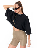 A woman modeling a black cropped t-shirt with wide three-quarter sleeves and a silver collar detail, paired with beige fitted biker shorts, against a plain white background.