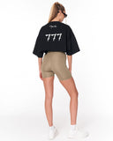 A woman shown from the back wearing a black oversized crop top with a white '777' graphic and 'Martin Valen' text, paired with beige fitted biker shorts and white sneakers, against a plain white studio background.