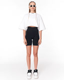 A model stands against a white studio background wearing a white cropped tunic top with wide draped sleeves, black fitted biker shorts, white platform sneakers, and white sunglasses.