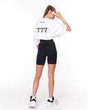 A woman shown from the back wearing a white cropped t-shirt with '777' printed in black on the back, black biker shorts, and white sneakers, against a plain white studio background.