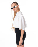 A woman modeling a white cropped tunic top with wide draped sleeves and black high-waisted shorts against a plain white background.