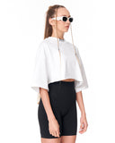 A model wearing a white cropped tunic top with wide draped sleeves, black fitted biker shorts, and white sunglasses with long chain details, shown against a plain white background.
