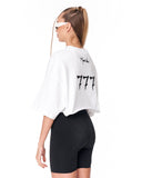 A woman shown from the back wearing an oversized white cropped T-shirt with black graphic text and black fitted biker shorts against a plain white studio background.