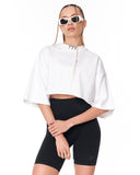 A model wearing a white cropped t-shirt with wide sleeves, black biker shorts, and white sunglasses with gold chains, photographed against a solid white background.