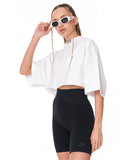 A model wearing a white cropped tunic top with wide sleeves, black high-waisted biker shorts, and white-framed sunglasses with chain straps, posed against a solid white background.