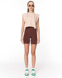 A model stands against a plain white studio background wearing a sleeveless light beige crop top, dark brown fitted biker shorts, white socks, and green-and-white sneakers.