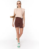 A woman modeling a sleeveless light beige crop top and dark brown bike shorts against a plain white studio background.