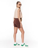 A woman modeling a sleeveless beige crop top, brown fitted biker shorts, white socks, and green and white sneakers, photographed against a plain white studio background.