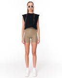 A model stands against a white studio background wearing a black sleeveless cropped top with a gold zipper, beige fitted biker shorts, white sneakers, and white-framed sunglasses.